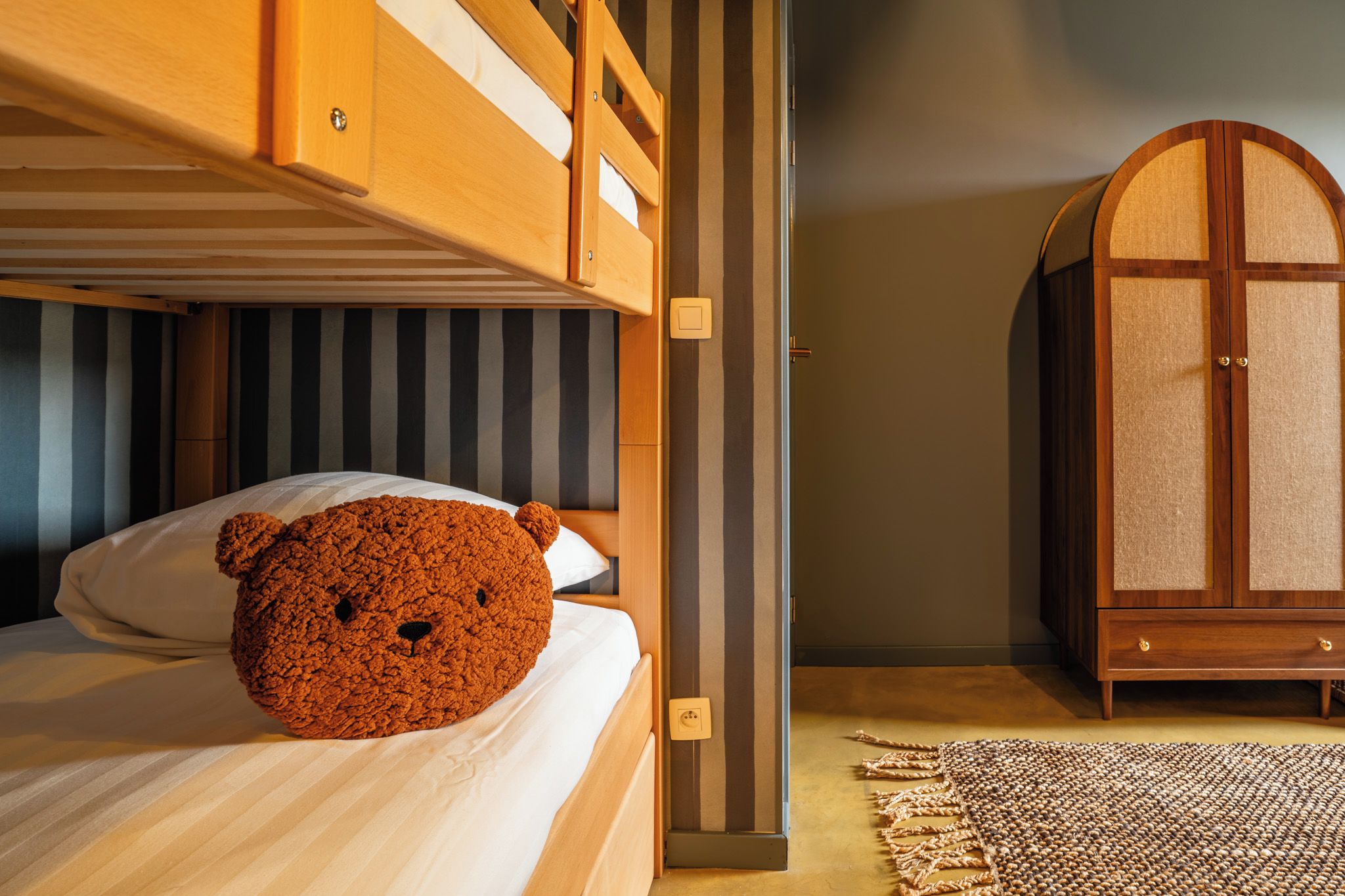Une chambre avec un lit superposé, un ours en peluche sur le lit du bas, une armoire et un tapis tissé.