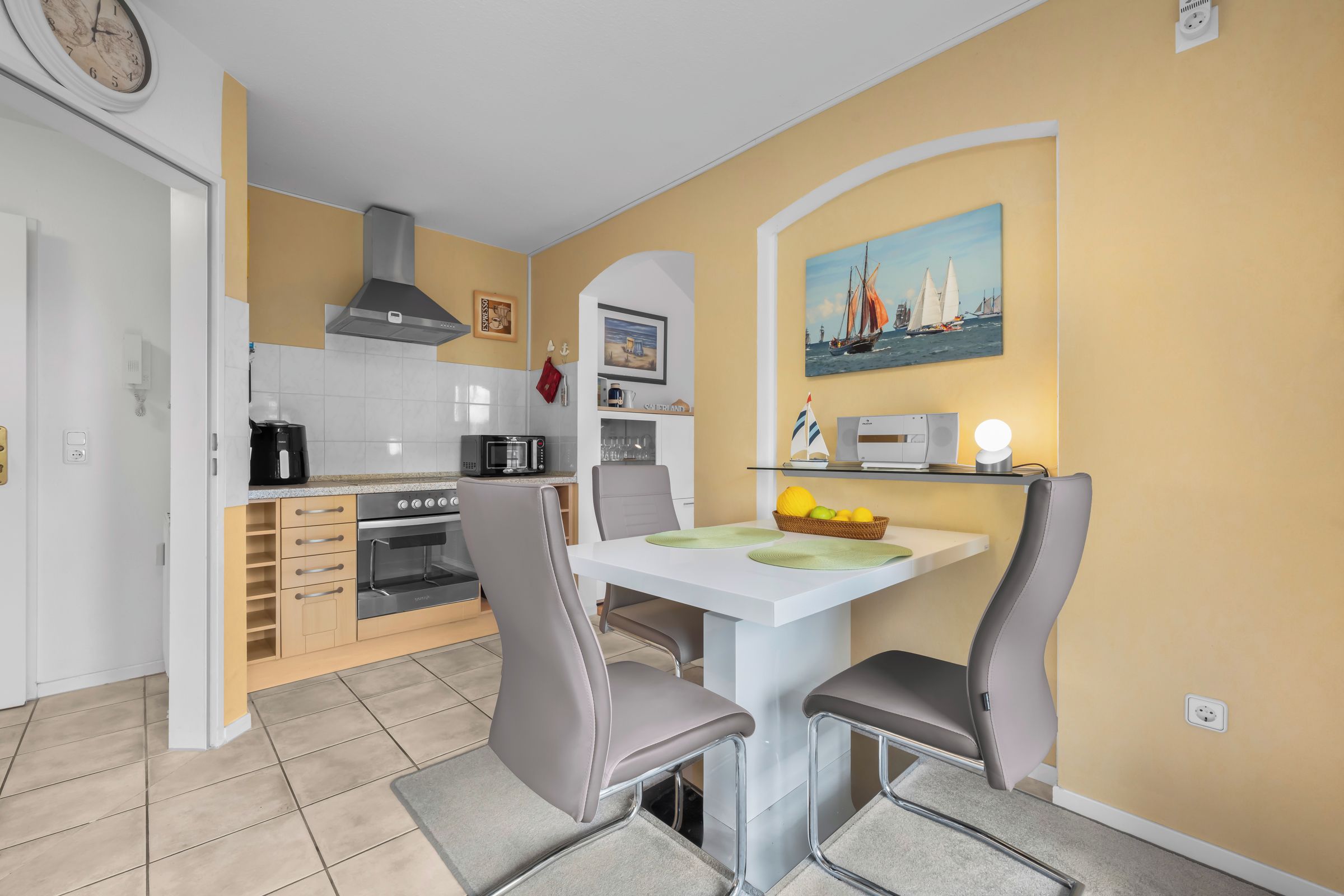 Een kleine keuken met een eethoek, met een magnetron, fornuis en een schilderij van zeilboten aan de muur.