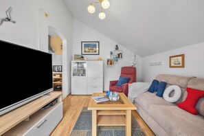 A modern living room with a flat-screen TV, a red armchair, a sofa with colorful cushions, a coffee table, and a bookshelf with various items. - Frische Brise Vacation Rental—Your Vacation Just 100 Meters from the Beach (Schönberg (Holstein))