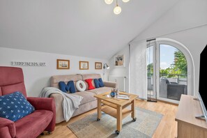 A modern living room with a red armchair, a beige sofa with colorful pillows, a wooden coffee table, and a large window with a view of greenery. - Frische Brise Vacation Rental—Your Vacation Just 100 Meters from the Beach (Schönberg (Holstein))