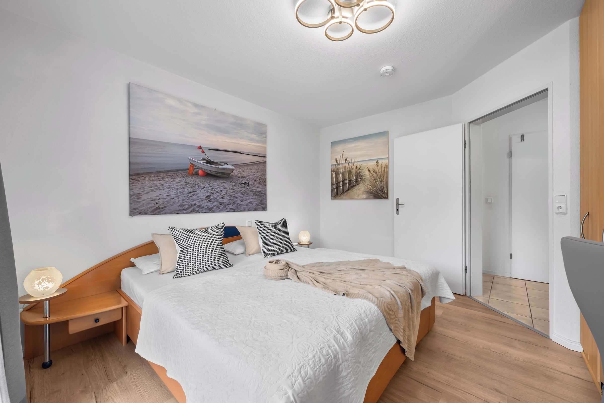 Un dormitorio con cama, mesitas de noche, una lámpara de pared y una obra de arte que representa una escena de playa.