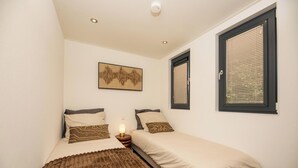 Une petite chambre bien éclairée, avec deux lits simples, une table de chevet avec une lampe, une œuvre d’art accrochée au mur et une fenêtre avec des stores.