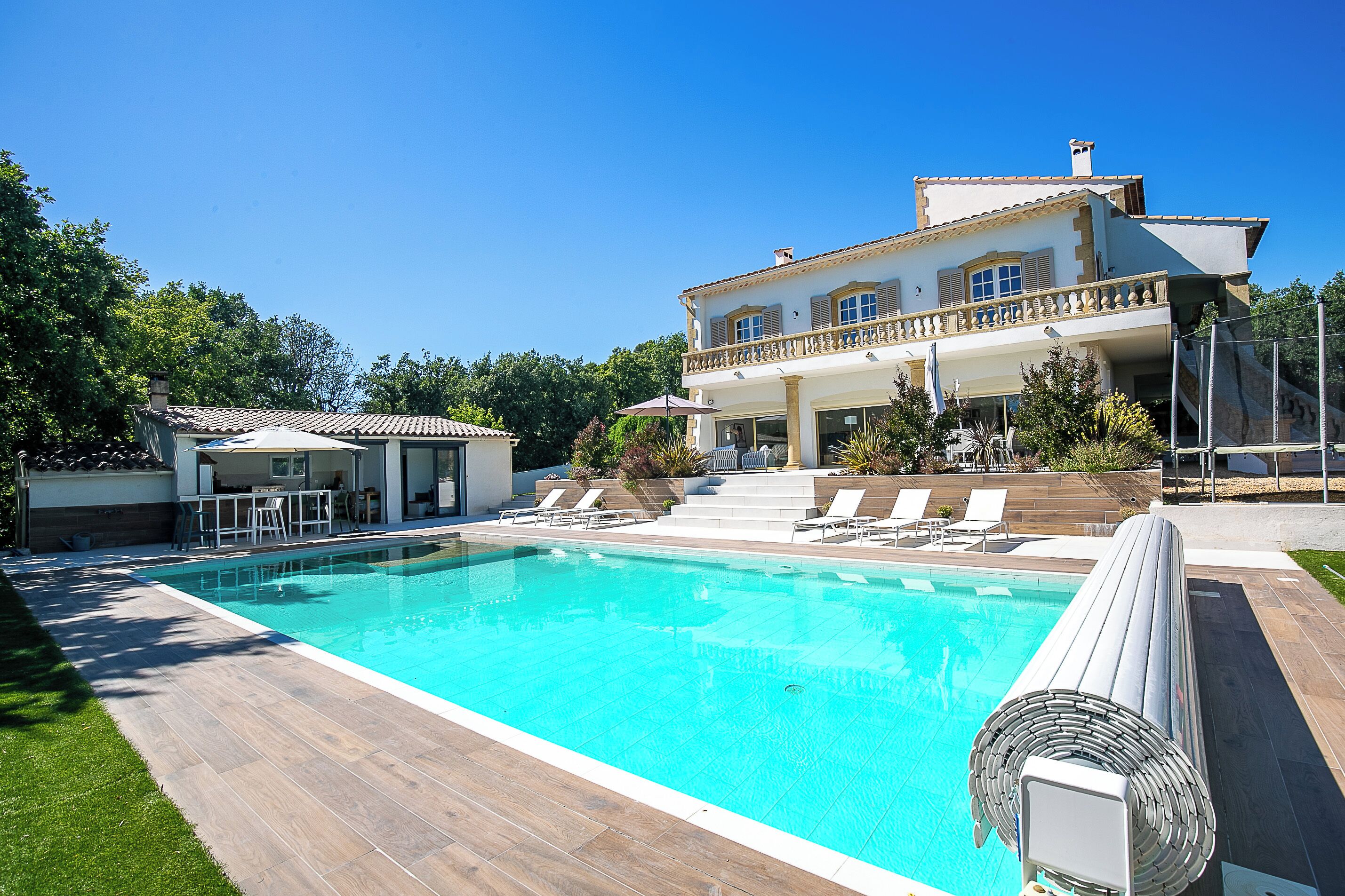 Une grande piscine avec une terrasse en bois, des chaises longues et un bâtiment à deux étages doté d’un balcon.