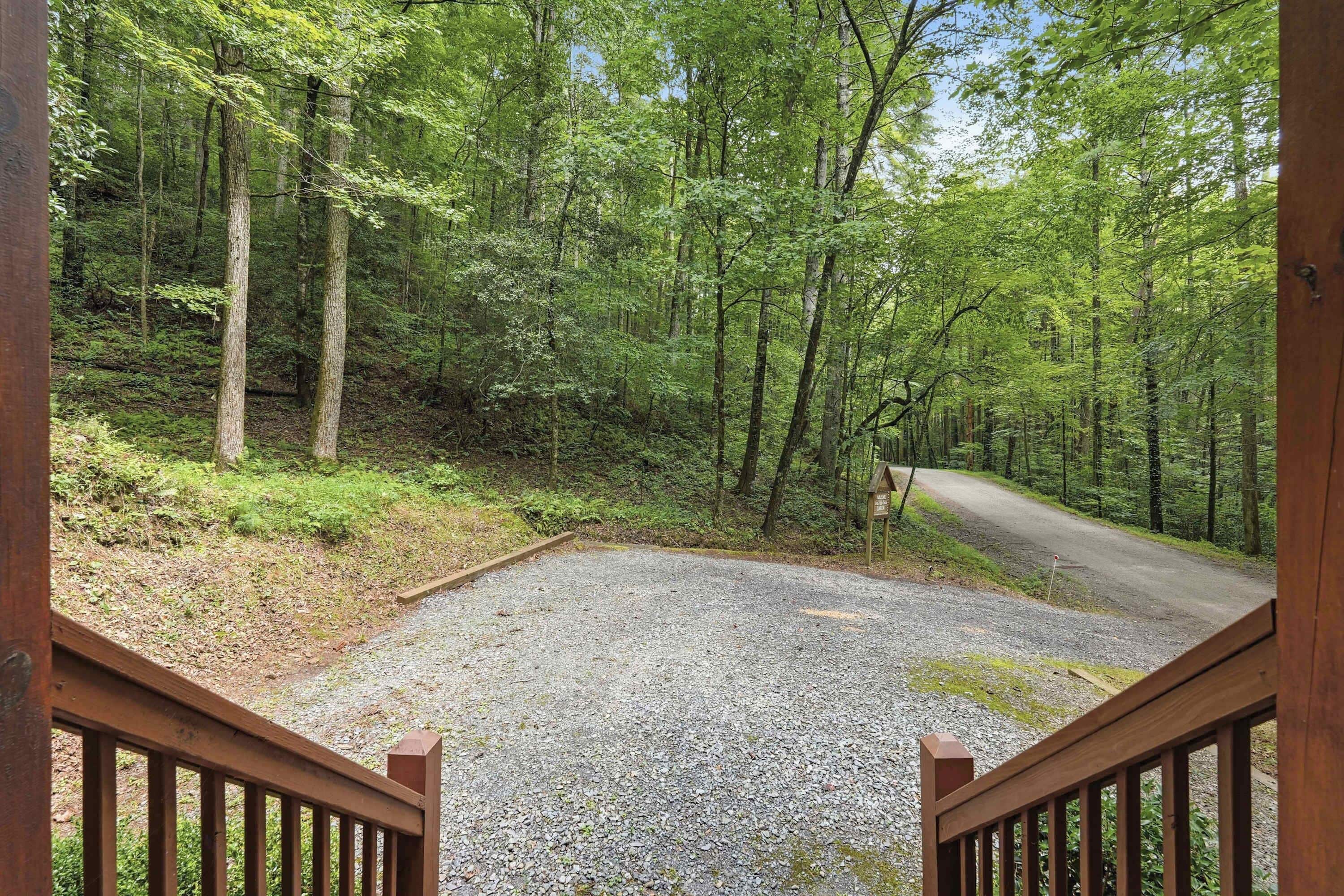 A gravel parking area leading to a forested path with a wooden fence and a signpost.