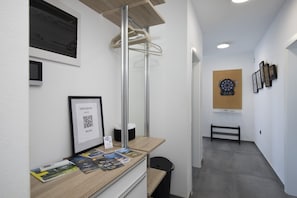 Uma mesa de recepção com um código QR emoldurado, uma pequena lixeira e um banco em um corredor com um alvo de dardos na parede.