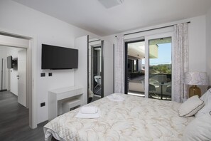 Ein modernes Hotelzimmer mit Flachbildfernseher, einem Bett mit gesteppter Bettdecke, einem Balkon mit Tisch und Stühlen und einer Schiebetür zum Balkon.