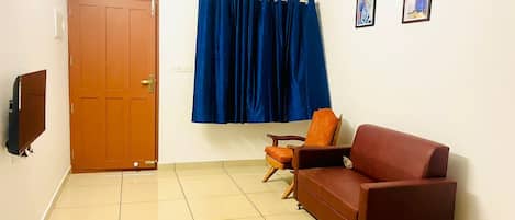 A room with a wooden door, a blue curtain, a brown sofa, a wooden chair, and a flat-screen TV mounted on the wall.