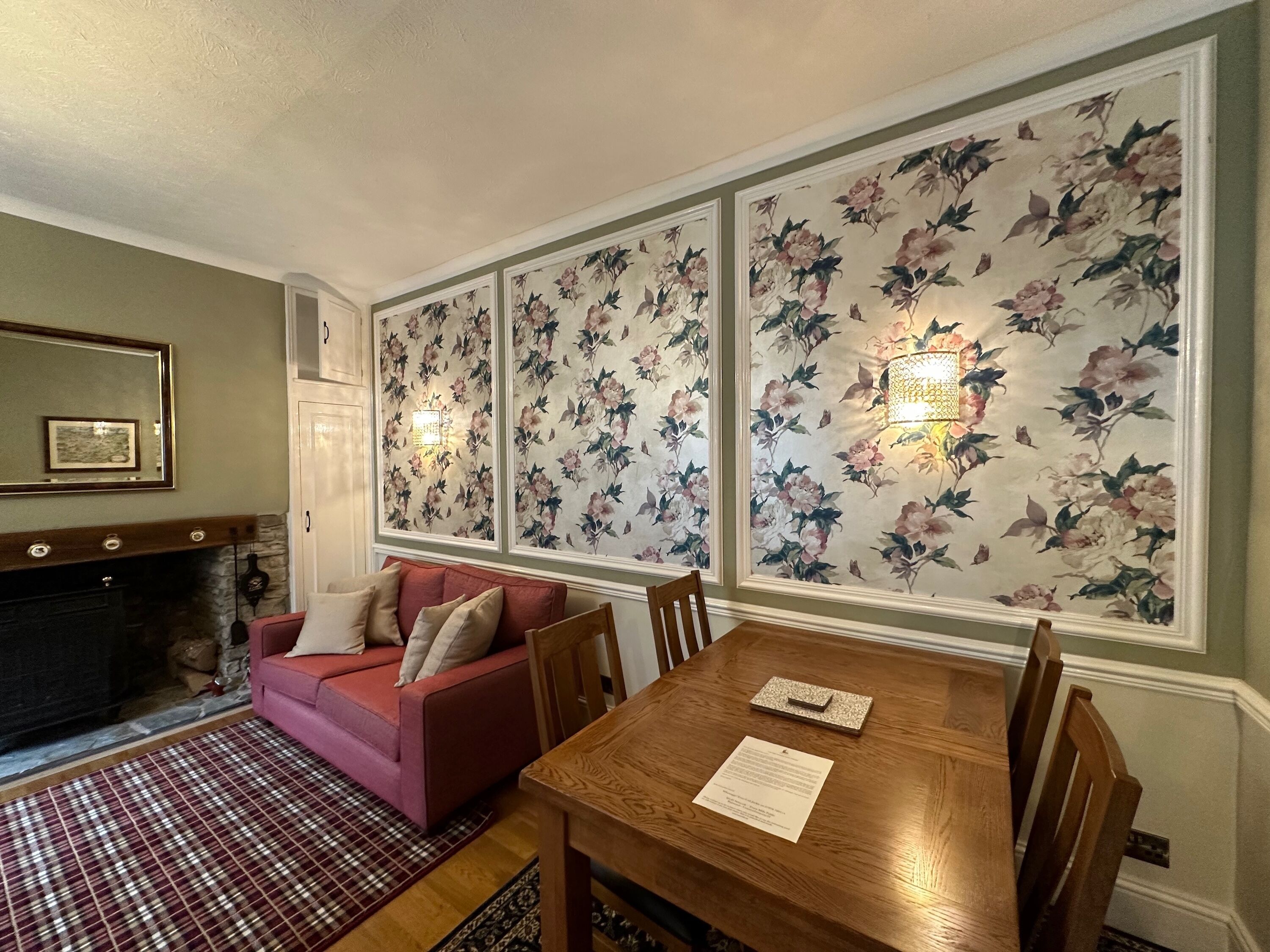 Una sala de estar con chimenea, un sofá rojo, una mesa de comedor de madera y papel tapiz floral.
