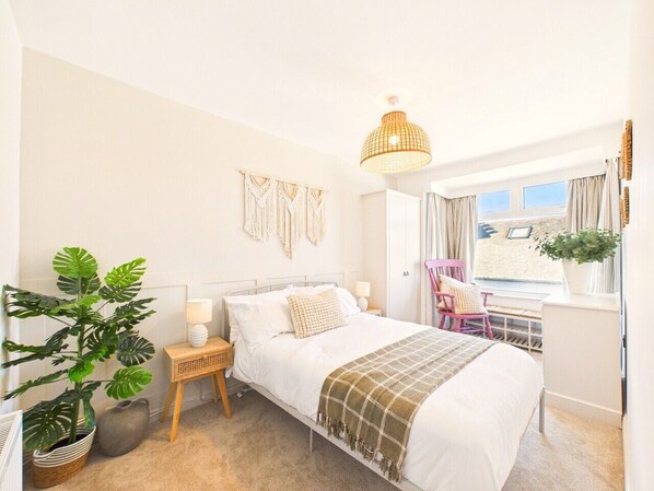 Une chambre à coucher avec un lit, des tables de chevet, un luminaire suspendu, une fenêtre avec des rideaux et une plante en pot.