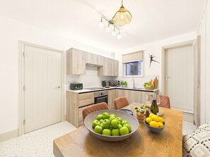 A modern kitchen with wooden cabinets, a dining table with a bowl of apples and lemons, and a pendant light. - Coastal Charm | 4BR House | near St Ives Harbour (St Ives)