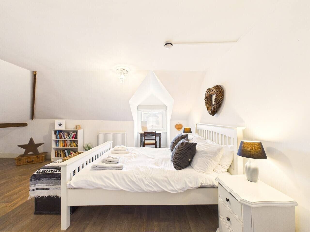 Un dormitorio con cama blanca, mesita de noche y lámpara. Hay una estantería y una decoración con forma de estrella en el suelo.