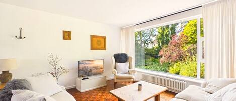 A modern living room with a sofa set, a coffee table, a television, and a large window with a view of greenery.