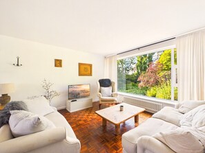 Ein modernes Wohnzimmer mit einer Sofagarnitur, einem Couchtisch, einem Fernseher und einem großen Fenster mit Blick auf Grünflächen.