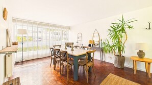 Un área de comedor con una mesa y sillas, un ventanal con persianas, una planta en maceta y una lámpara sobre una mesita.