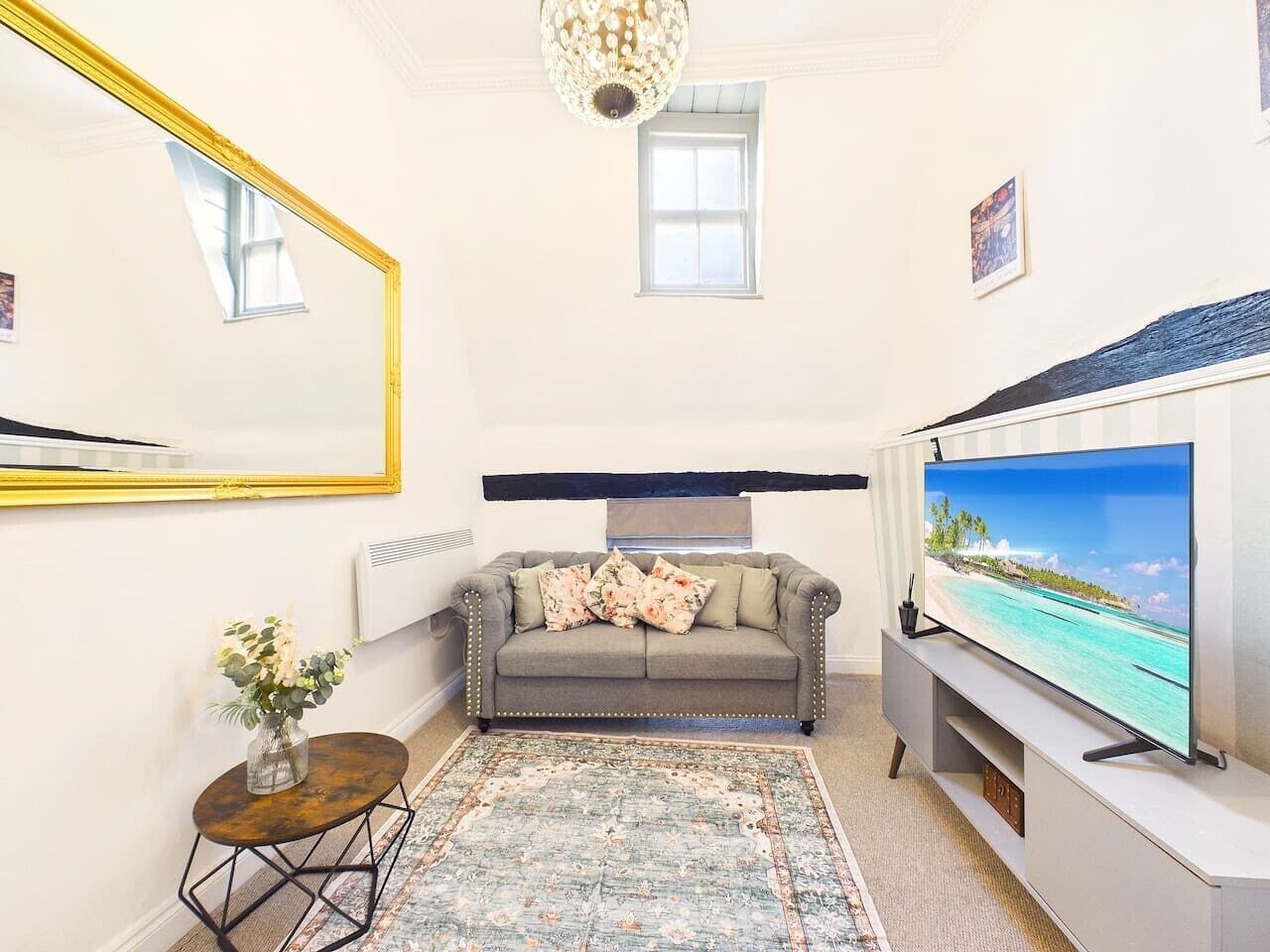 A living room with a sofa, a television, a rug, a small table with a vase, and a large mirror.