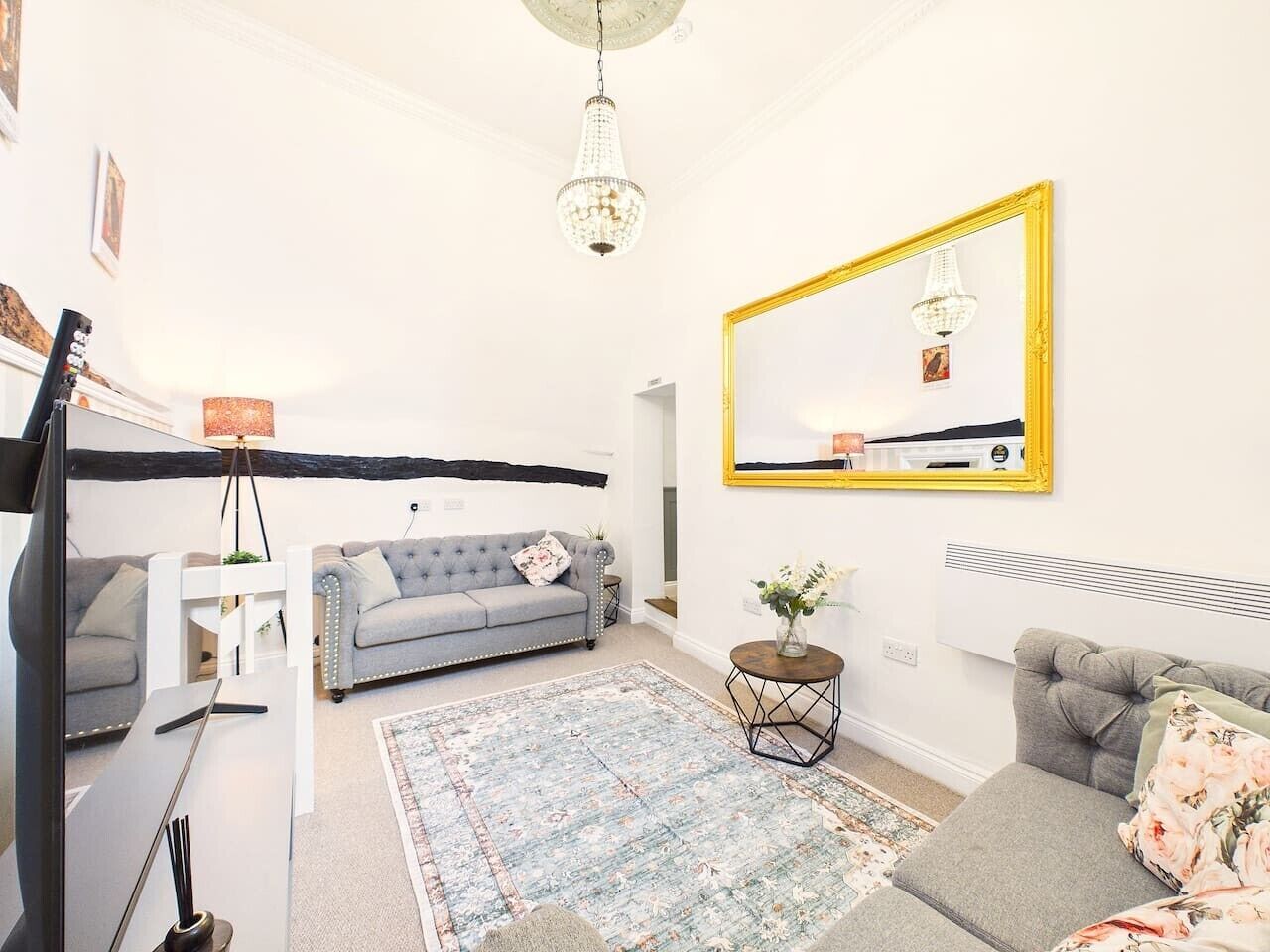 A living room with a grey sofa, a round coffee table, a chandelier, and a large mirror with a yellow frame.