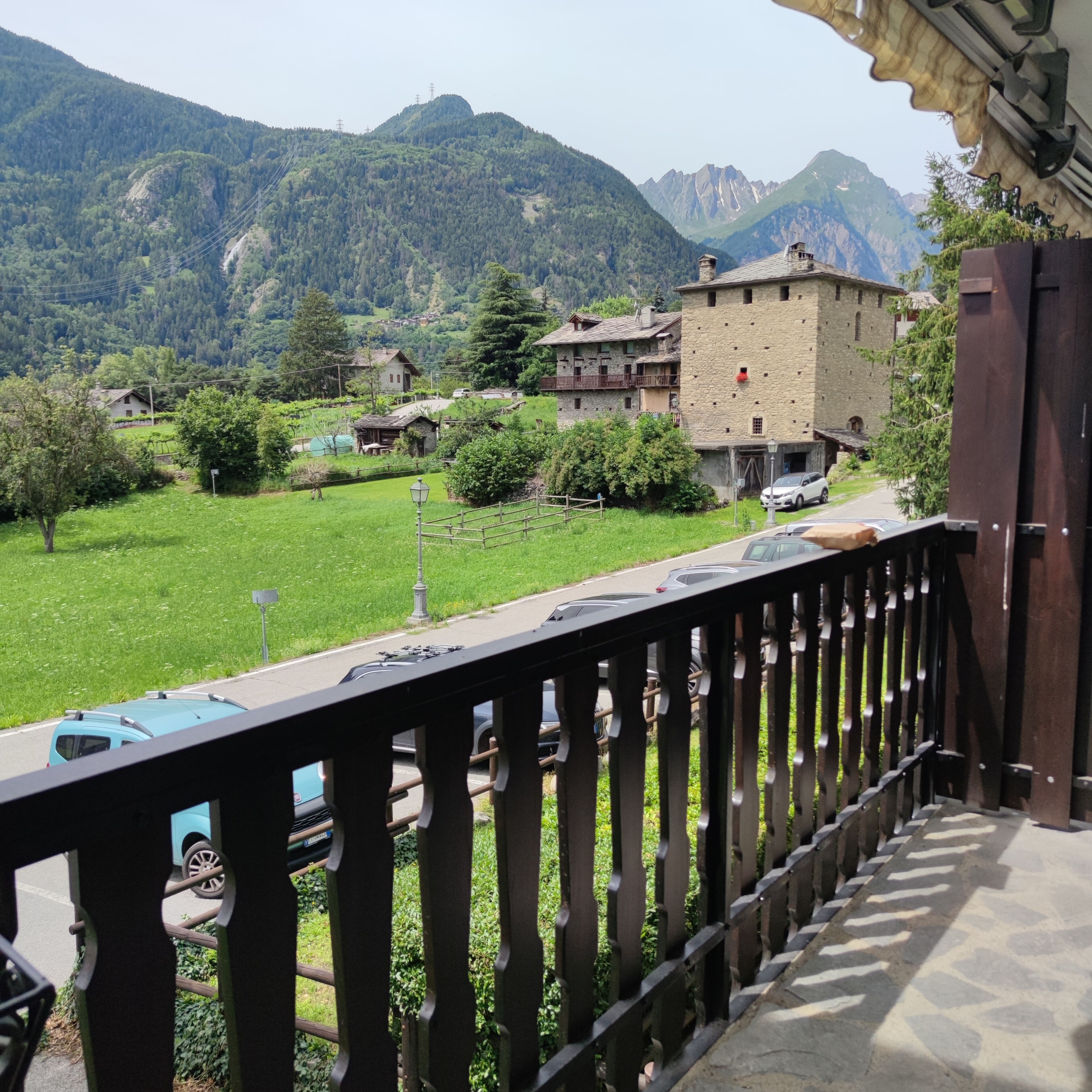 Ein Balkon mit Blick auf eine Berglandschaft, ein Steinhaus und abgestellte Autos.