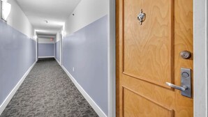 A hotel hallway with a carpeted floor, a wooden door with a lock, and a light fixture on the wall. - Chic City Condo With Pool & Gym (Miami)