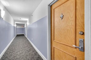 A hotel hallway with a carpeted floor, a wooden door with a lock, and a light fixture on the wall. - Chic City Condo With Pool & Gym (Miami)