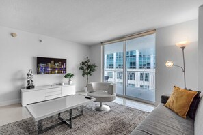 A modern living room with a white sofa, a white coffee table, a grey rug, a white cabinet, a plant, and a cityscape painting on the wall. - Chic City Condo With Pool & Gym (Miami)