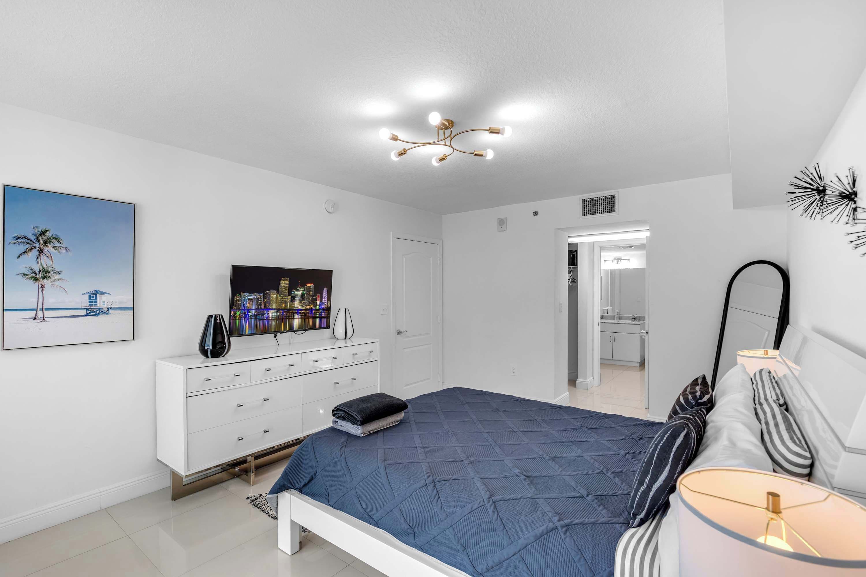 Une chambre à coucher comprenant un lit, une commode, un téléviseur et un tableau représentant une scène de plage.