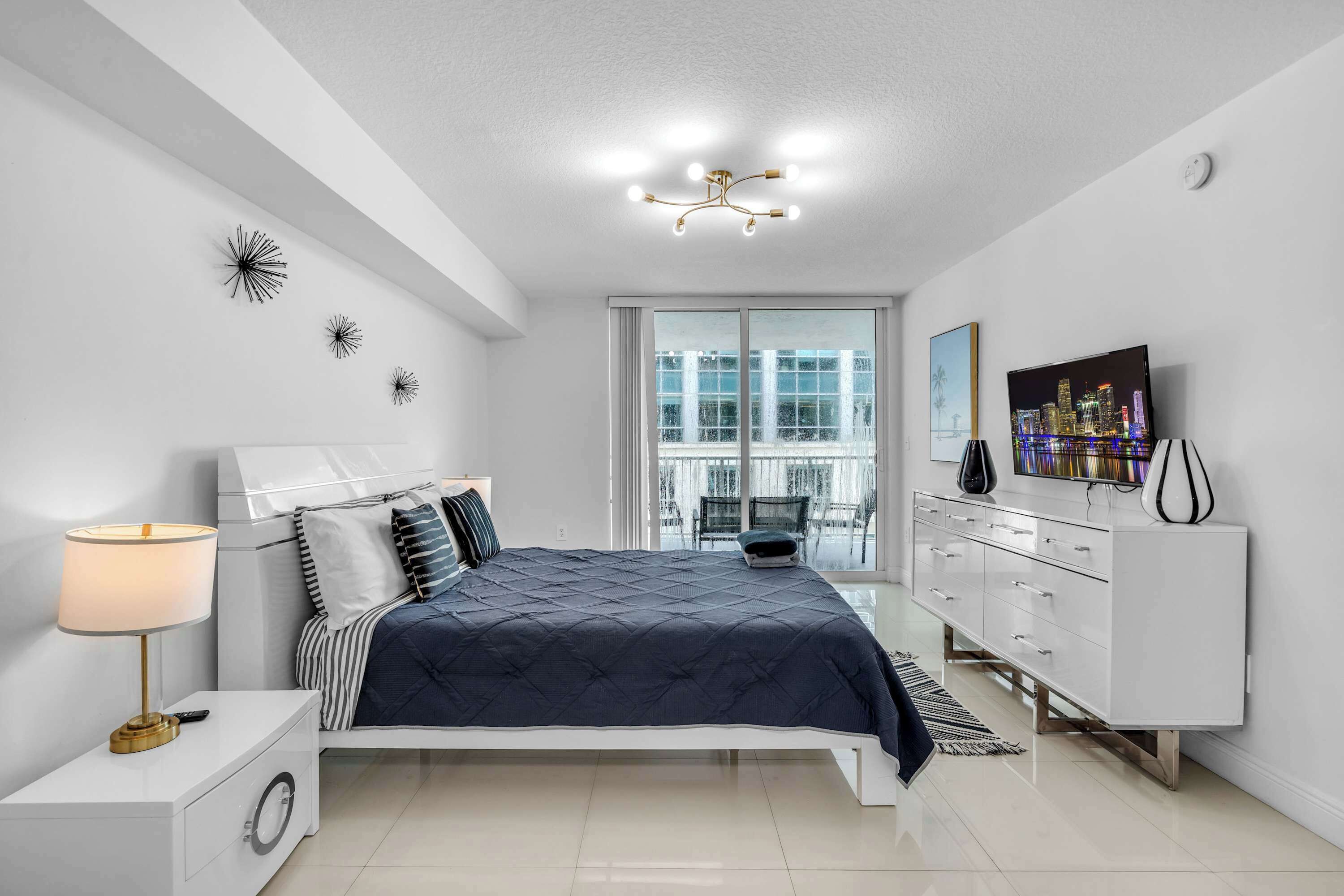A modern bedroom with a bed, bedside table, lamp, dresser, and a cityscape view.