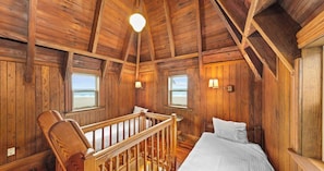 Une chambre de cabane en bois avec un lit, deux fenêtres donnant sur la plage, une rambarde en bois et un luminaire suspendu.