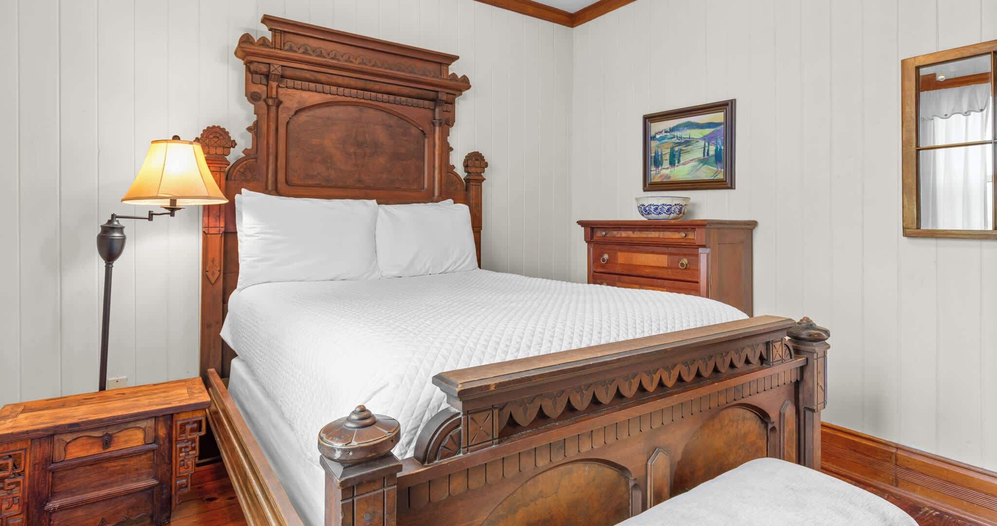 Een klassiek houten bed met hoofdeinde, nachtkastje, lamp en een schilderij aan de muur.