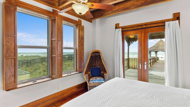 Une chambre avec un lit, un fauteuil en osier et une vue sur la plage, grâce à de grandes fenêtres.