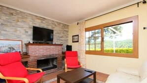 A living room with a brick fireplace, a television mounted above it, two red chairs, a white sofa, and a window with a view of greenery.
