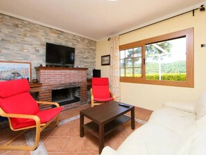A living room with a brick fireplace, a television mounted above it, two red chairs, a white sofa, and a window with a view of greenery.