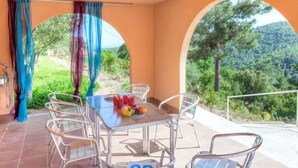 A covered outdoor dining area with a table and chairs, a view of greenery and trees, and a clear sky.