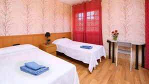 A hotel room with two single beds, a desk, a chair, a nightstand, a lamp, and a ceiling fan.