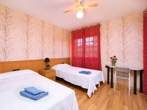 A hotel room with two single beds, a desk, a chair, a nightstand, a lamp, and a ceiling fan.