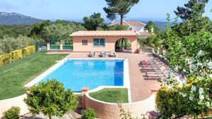 A poolside area with a clear view of the pool, a small building, and a hilly landscape in the background.