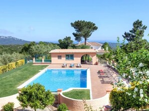 A poolside area with a clear view of the pool, a small building, and a hilly landscape in the background.