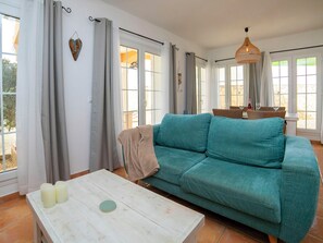Ein Wohnzimmer mit einem petrolfarbenen Sofa, einem weißen Couchtisch mit Kerzen und einem Essbereich mit Korbmöbeln.