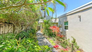 A narrow pathway flanked by lush greenery and a wooden fence, leading to a building with a window and a door. - Ventur Escapade Heated Pool Guesthouse (Wilton Manors)