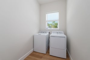 A laundry room with two white stacked washer and dryer units, a window with blinds, and a wooden floor. - Seashell Oasis - Heated Pool/Hot Tub/Putting Green/Walk to Beach (St. Augustine)