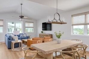 A modern living room with a blue sofa, orange chairs, a wooden dining table, and a ceiling fan. - Seashell Oasis - Heated Pool/Hot Tub/Putting Green/Walk to Beach (St. Augustine)