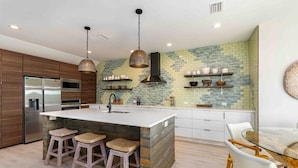 Et moderne køkken med en central ø, barstole, rustfrie hårde hvidevarer og et backsplash med et geometrisk mønster.