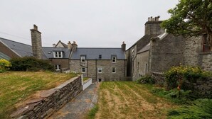 Un bâtiment en pierre avec une entrée centrale, entouré d’un mur de pierre et d’une zone herbeuse.