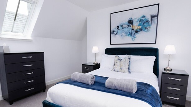 Une chambre à coucher comprenant un lit, deux tables de chevet, une commode, une fenêtre et un tableau abstrait accroché au mur.