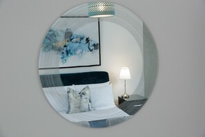 A circular mirror reflecting a bed with a white headboard, a pillow, and a bedside lamp.