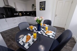 A modern kitchen with white cabinets, a black countertop, and a dining area with a checkered table mat, chairs, and a vase of flowers. - Central Lerwick House, Modern, 2 Bathrooms, Garden (Lerwick)