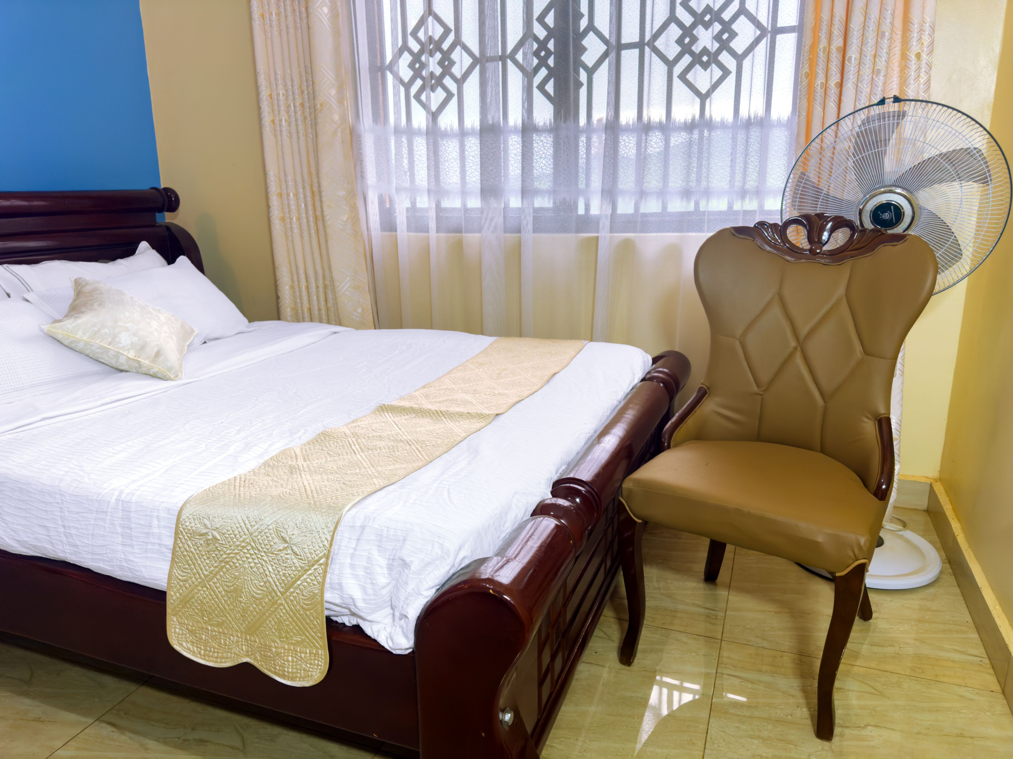 A neatly made bed with a wooden headboard, a beige chair with a fan, and a window with patterned curtains.