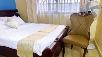 A neatly made bed with a wooden headboard, a beige chair with a fan, and a window with patterned curtains.
