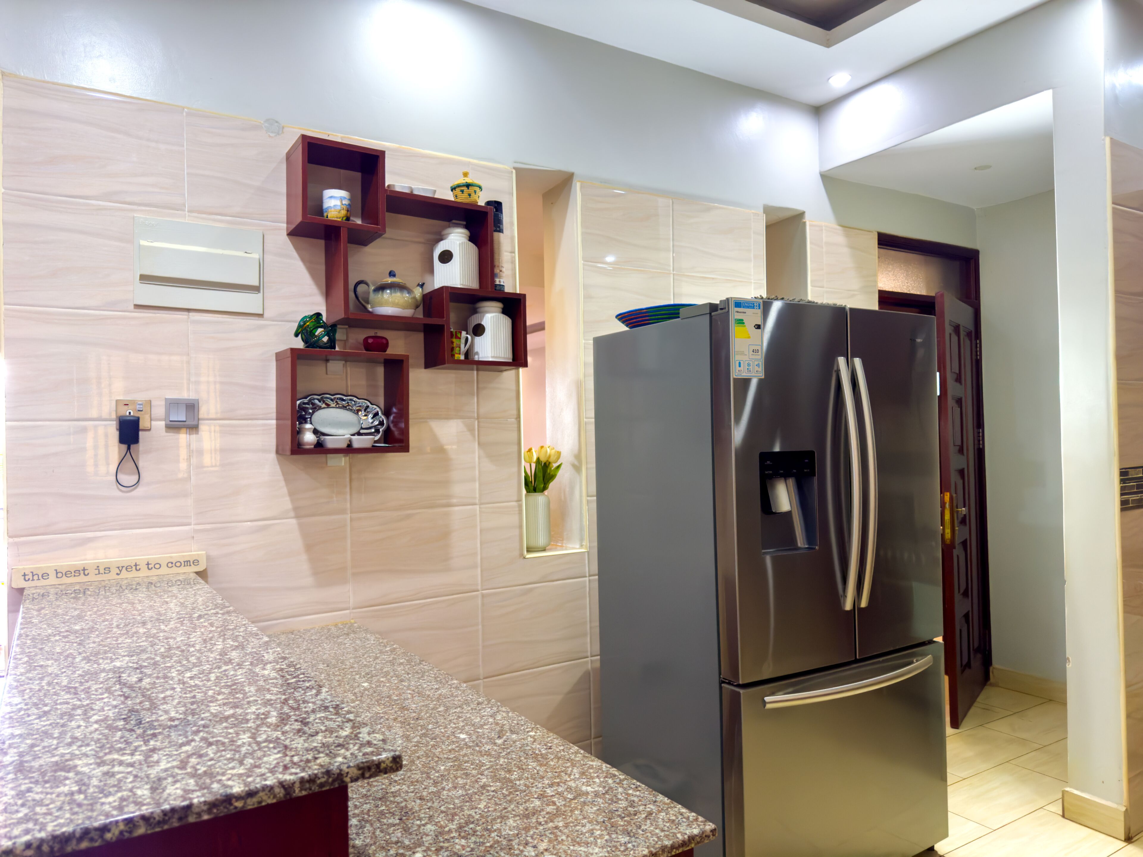 A modern kitchen with a stainless steel refrigerator, granite countertop, and wooden shelves with kitchenware.