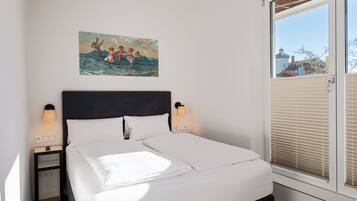 Ein ordentlich bezogenes Bett mit weißen Leinen, ein dunkles Kopfteil und zwei Nachttischlampen. Über dem Bett hängt ein Gemälde einer Strandansicht.