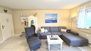 Ett vardagsrum med en grå soffa, en tillhörande fåtölj, ett vitt soffbord och en tavla med en segelbåt på väggen.